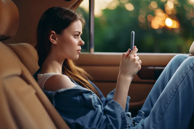 Young woman using smartphone in a car during sunset, wearing casual denim outfit and reflecting a serene mood with a soft background.