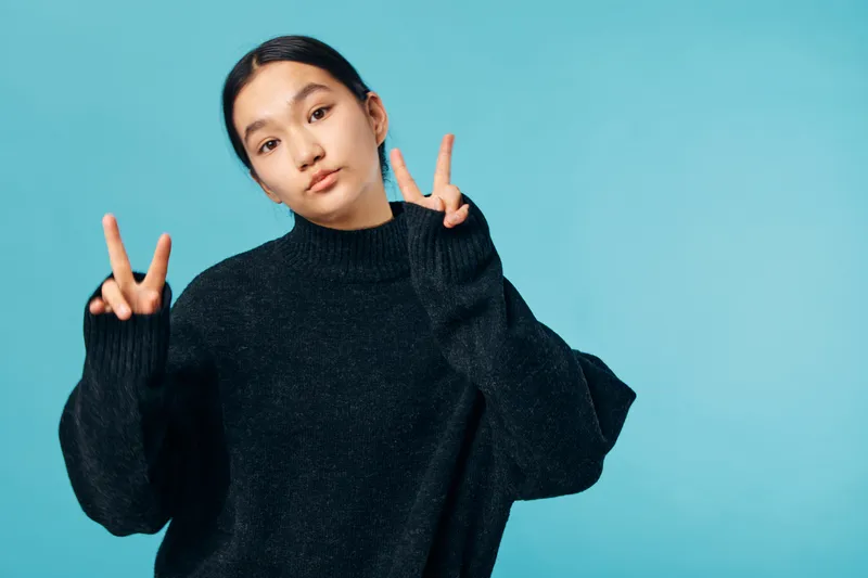 Young woman wearing black sweater showing peace sign with both hands looking at camera against blue background casual style and confident expression in studio portrait.