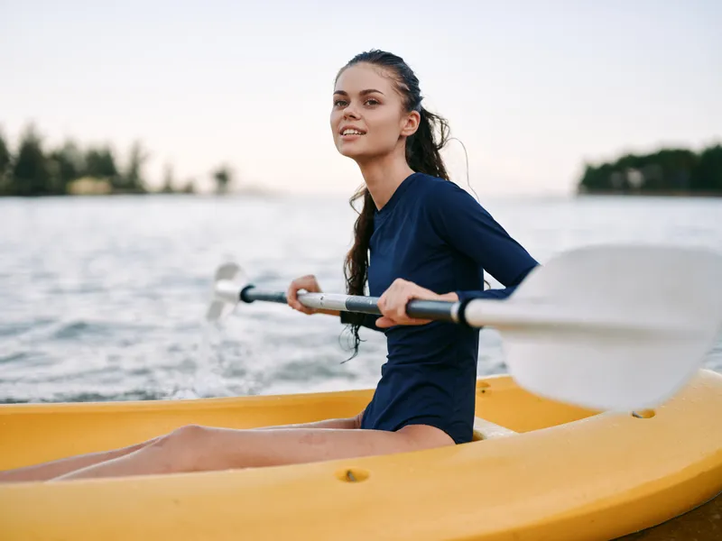 An athletic woman sits in a yellow kayak on calm water, paddling with a double blade paddle, smiling toward the camera and enjoying a sunny outdoor moment.