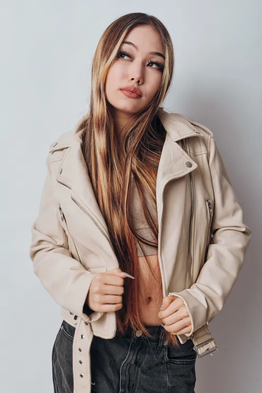 Young woman with long hair wearing beige leather jacket and crop top posing against light background in studio. Fashion style, casual outfit and confident expression for portrait.