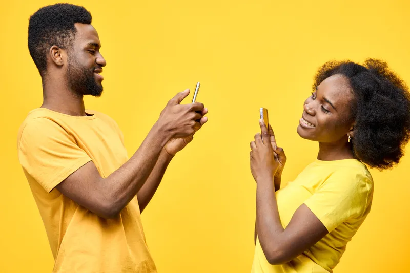 A cheerful young African couple joyfully engaging with their smartphones, set against a vibrant yellow background, showcasing contemporary casual fashion