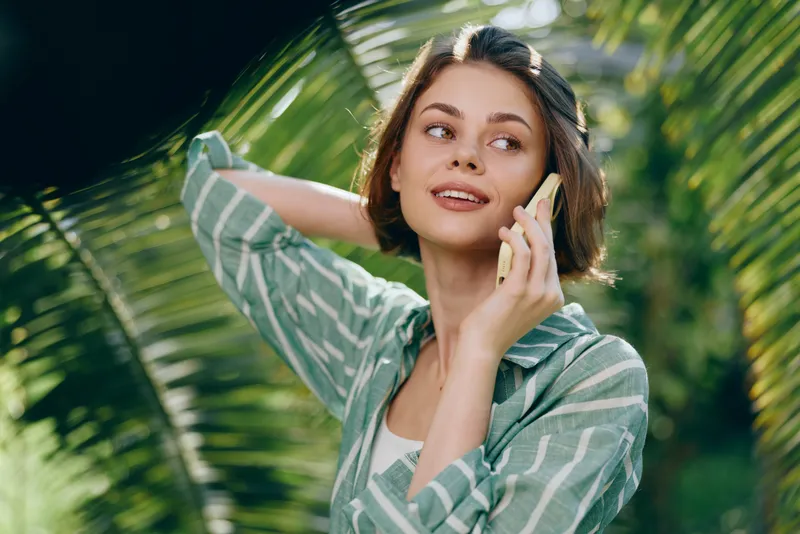 Woman outdoors, smiling, talking on a phone amid lush greenery. Casual striped shirt, relaxed pose, sunny day, natural light, cheerful mood, everyday moment, and warm vibe for creative use.