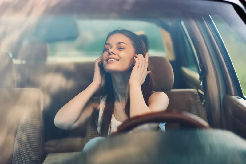 Young woman enjoying music in a car, smiling and carefree on a sunny day, reflecting happiness and relaxation while driving in a suburban area.