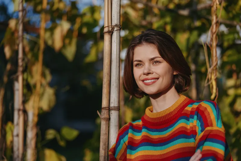 Woman portrait smile sweater colorful outdoors garden sunlight, young female in striped knitwear smiling in golden hour. Cozy casual fashion, relaxed cheerful mood and warm autumn foliage.