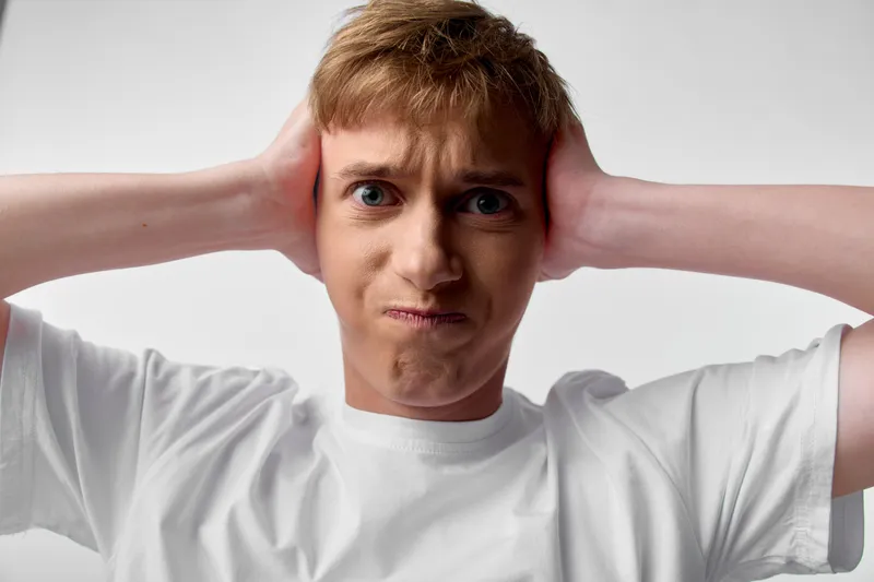 Man holds his ears with both hands, showing frustration at loud noise in a plain studio, capturing discomfort and the urge to block out sound in a simple, focused image