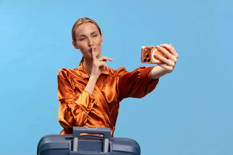 Blonde woman in shiny orange blouse takes selfie with smartphone, holding finger to lips in shushing gesture, standing behind blue suitcase on blue background. Concept of travel and secrecy.