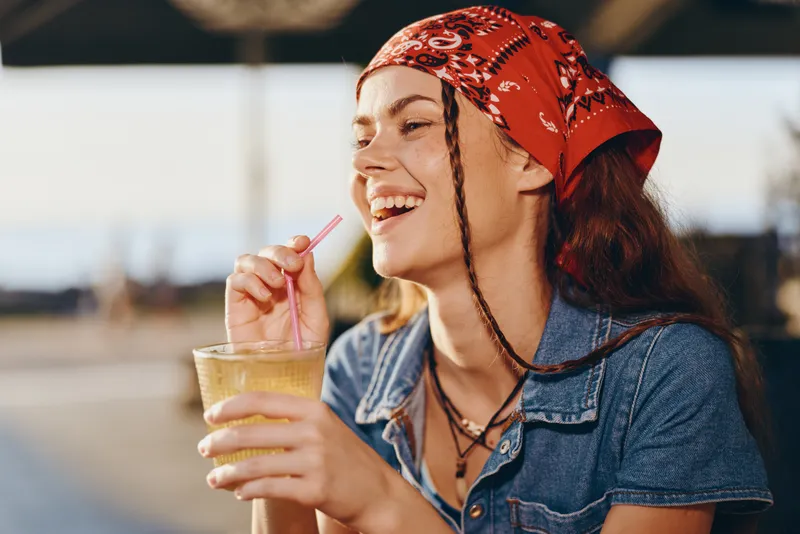 Lifestyle woman in modern boho-western denim street style enjoying a drink with a joyful smile, film color tones capturing casual summer vibes and relaxed mood.