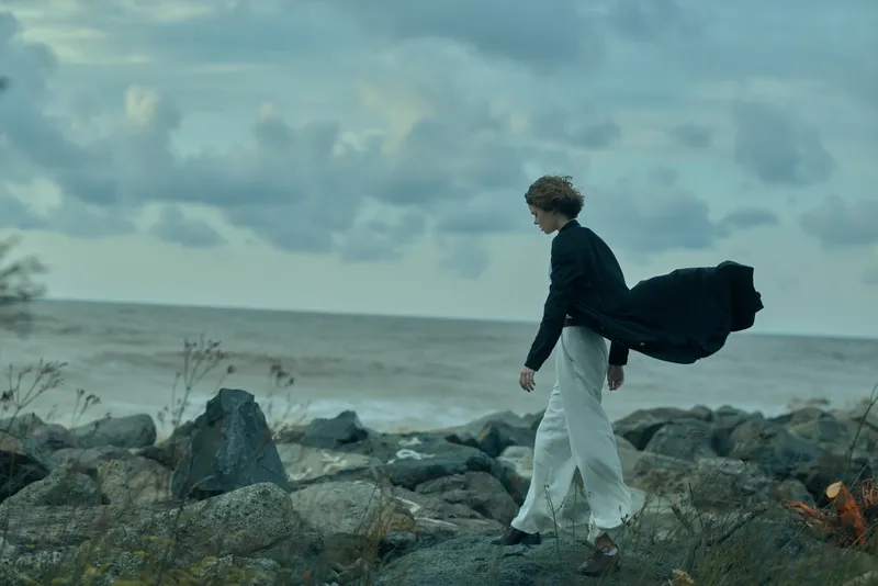 Woman walks along a rocky shoreline with a billowing coat, cloudy sky and distant horizon, capturing moody movement and solitary exploration by the sea.