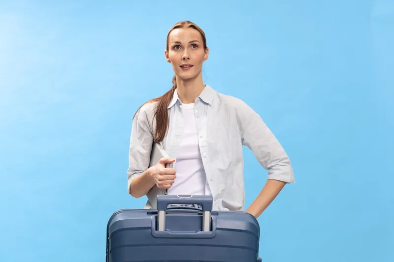 Young woman with long hair stands holding blue suitcase on blue background. Casual travel concept with relaxed confident female traveler ready for journey or vacation.