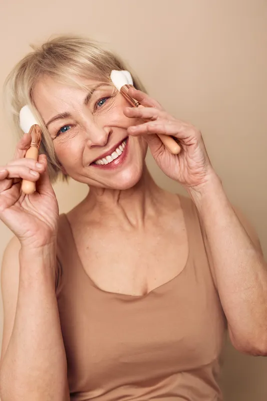 Smiling mature woman with short blonde hair applies natural skincare cream with cotton pads on face. Concept of beauty, self-care, wellness, and healthy skin routine on beige background.
