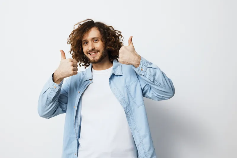 Happy young man with curly hair giving a thumbs up against a light background, wearing a casual light blue shirt, representing positivity and confidence