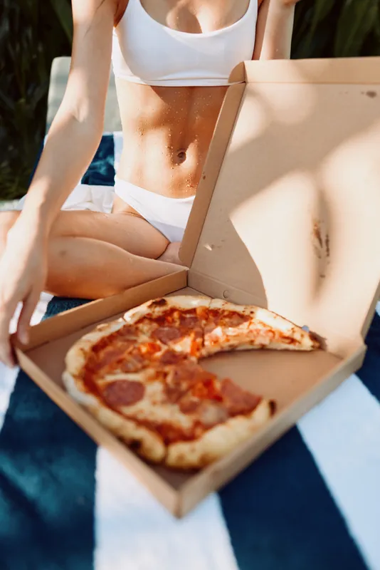 Cheerful woman outdoors, wearing white bikini, enjoying pizza from an open box on a colorful towel, sunny day, relaxed mood, casual meal, vibrant scene, summertime leisure and sunshine highlights,