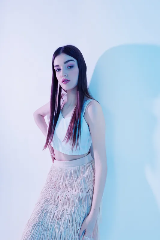 Young woman with long dark hair poses confidently in a white top and textured skirt against a soft blue background. Concept of beauty fashion and style.