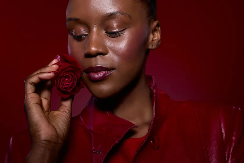 Close-up of a dark skinned African woman in red leather jacket holding a red rose near her face with closed eyes. Concept for beauty, elegance, and floral fragrance on red background.