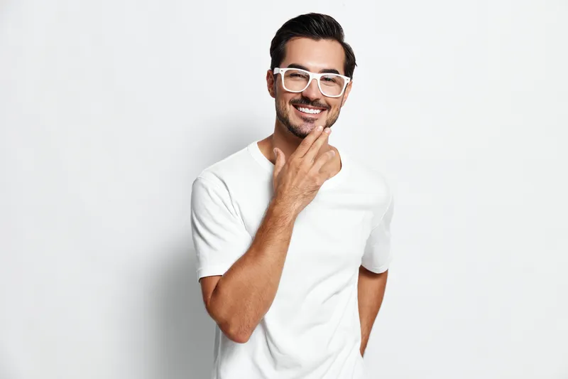 Smiling man in casual white t-shirt with stylish glasses poses cheerfully while touching his chin, conveying a sense of confidence and approachability against a bright background.