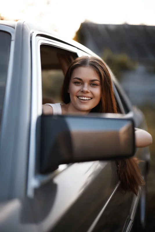 Happy young woman with long hair leaning out of a car window, enjoying a carefree moment on a sunny day, representing freedom and joy in travel.