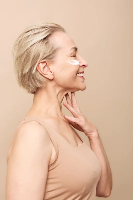 Mature woman with short blonde hair applies white face cream on cheek, smiling gently with eyes closed. Nude background and tank top create a soft, natural skincare and self-care concept.