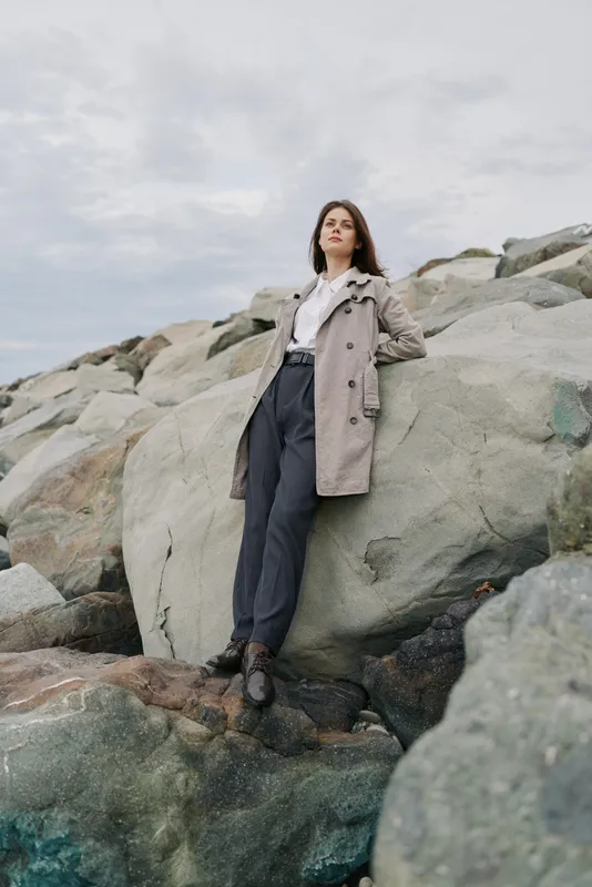 Young woman in beige trench coat and black pants stands confidently on rocks under cloudy sky. Concept of fashion, autumn, confidence, and outdoor lifestyle portrait.