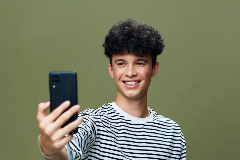 smiling man taking selfie with smartphone, casual striped shirt, curly hair, indoors with olive green background, cheerful young adult portrait