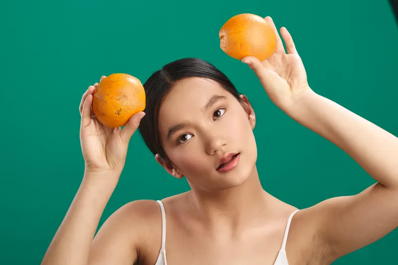 Asian woman playful portrait holds oranges near her head against a vibrant green backdrop, exuding fun, creativity, and a lighthearted mood in a bold studio setting.