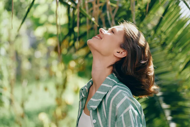 A woman outdoors, basking in sunlight, eyes closed and head tilted upward, wearing a striped shirt amid lush greenery.
