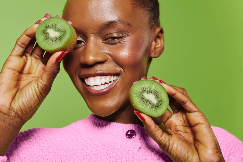 Smiling dark skinned African woman holds two halves of fresh kiwi fruit near face on green background. Concept of healthy lifestyle, fresh fruit, natural beauty, and wellness.