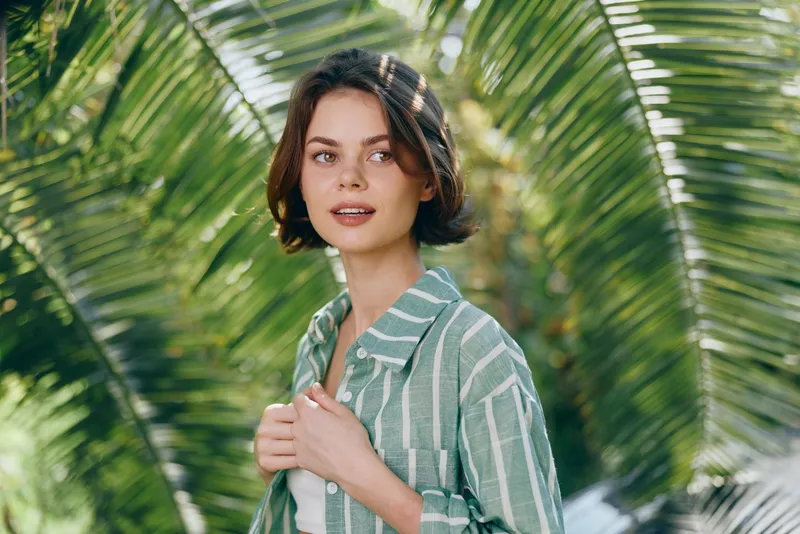 Portrait of a woman outdoors among tropical palms, wearing a striped green shirt, soft sunlight and a calm, natural expression.