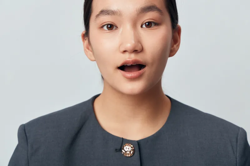 Young woman in formal suit with surprised expression on face against plain background. Closeup portrait of emotional female showing open mouth and raised eyebrows in studio.