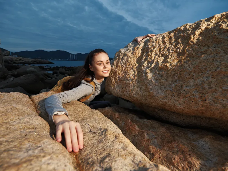 A young woman embracing a rocky landscape while smiling joyfully, showcasing adventure and outdoor exploration with a seaside backdrop