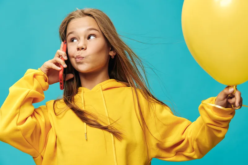Girl with yellow balloon and hoodie talking on smartphone against blue background looking thoughtful and playful with flowing hair and casual style in studio.