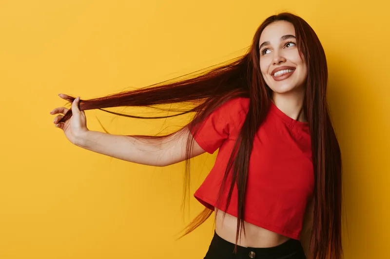 Young woman with long brown hair wearing red crop top smiling and holding hair strands on yellow background. Casual style, happy expression and playful mood with bright colors.