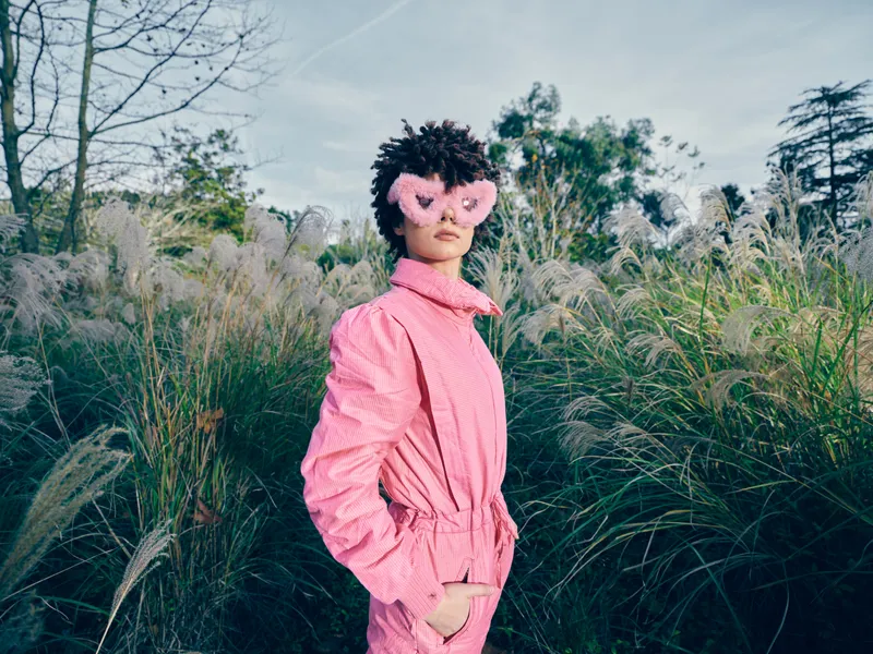 Fashion portrait of a model in a pink coat wearing a heart shaped pink mask in tall grasses, outdoor garden setting, playful glamorous style and whimsical mood.