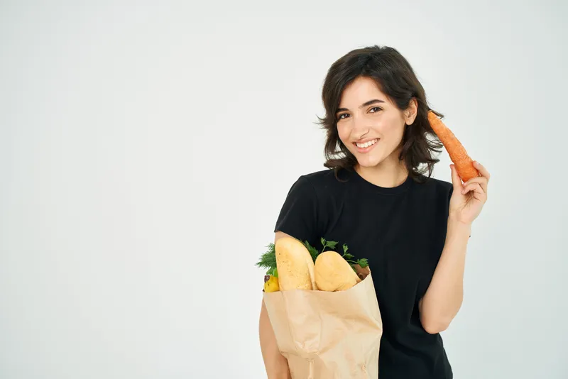 Весёлая брюнетка в чёрной футболке держит в руках пакет с продуктами, морковь и другие овощи