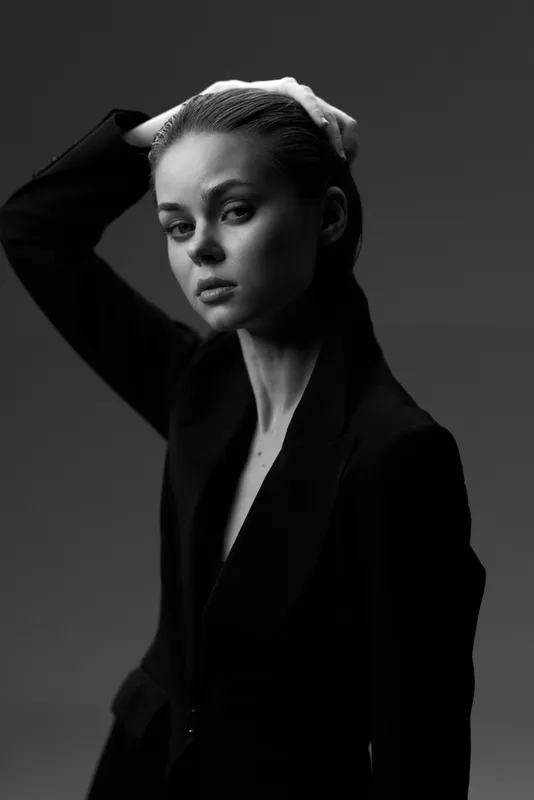 Black and white portrait of a serious young woman in a dark blazer with hand on head, expressing confidence and elegance.