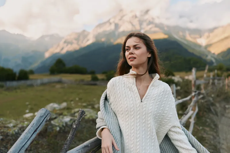 Young woman with long hair leans on a wooden fence in a mountain valley, wearing a white knit sweater and gazing into the distance with a serene, thoughtful expression.