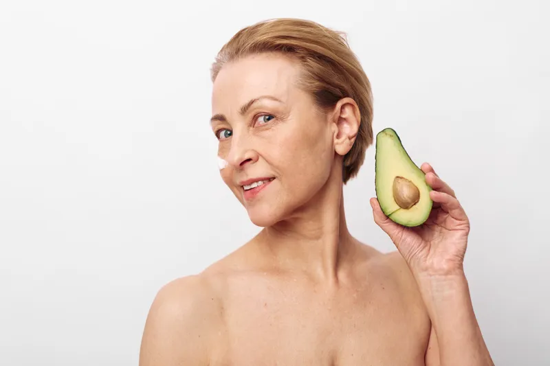 Mature woman with short hair holds half of avocado near face, smiling softly on white background. Concept of natural skincare, healthy lifestyle, and organic beauty treatment.