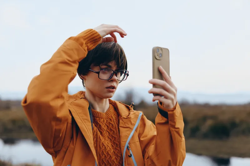 Woman selfie outdoors in autumn orange jacket adjusting hair while holding phone camera, lifestyle travel vibe, soft daylight and distant hills background, relaxed candid mood with soft warm breeze