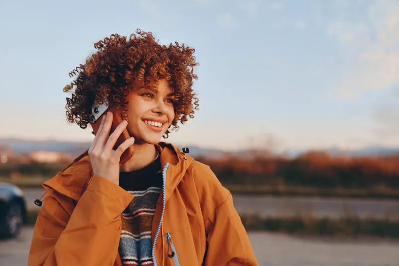 Woman curly hair smiles while making a smartphone call outdoors at sunset, wearing orange jacket, relaxed lifestyle, river background, shallow depth of field, evening portrait