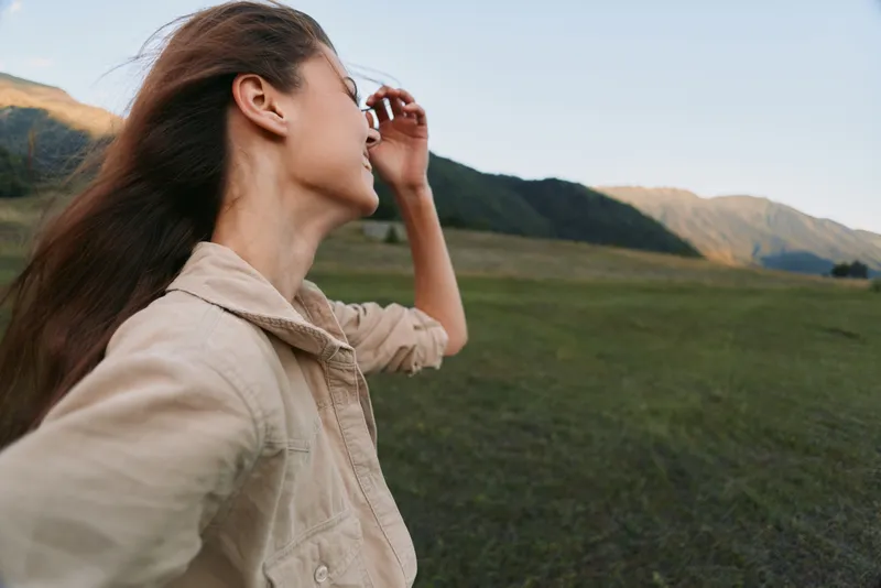 Young woman in an open field with distant mountains shields her eyes from the wind, enjoying a free-spirited moment in a casual beige jacket under soft light.