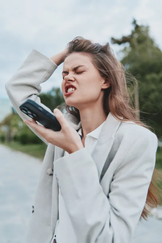 Woman in business attire expressing frustration while holding a phone outdoors, surrounded by nature, showcasing a modern, professional look.