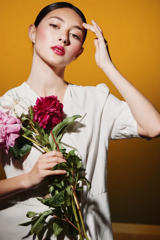 Young woman in white holds flowers close while posing against a bright background. Creative beauty and fashion concept with glossy lips and smooth skin, showcasing soft lighting.