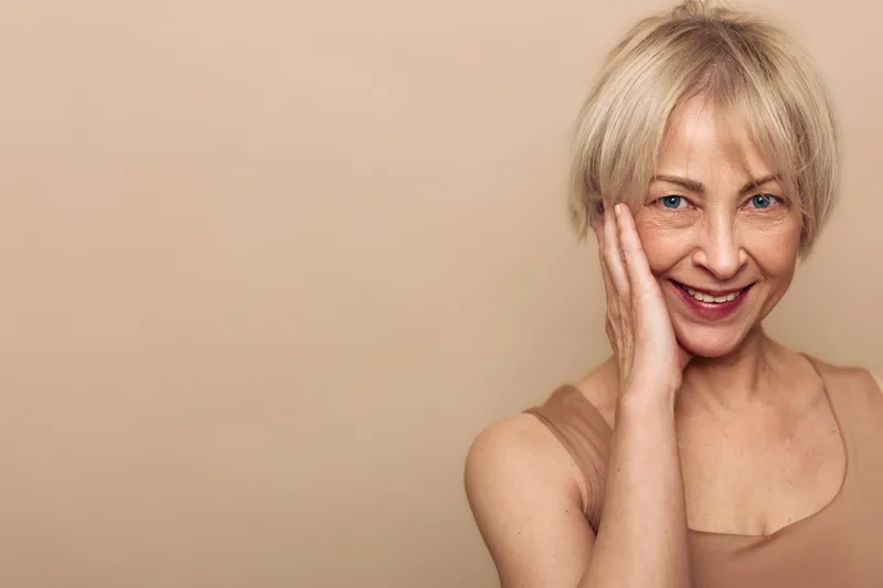 Smiling mature woman with short blonde hair touches her face gently, showing natural skin and happiness on beige background. Concept of beauty, aging, skincare, and wellness.