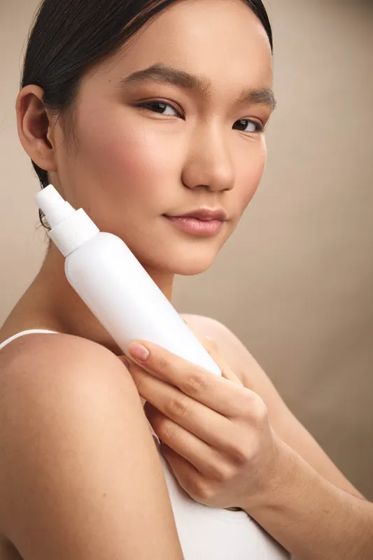 Asian woman holds a skincare bottle in a close view, presenting a calm, intimate moment. She smiles softly as she prepares to apply lotion, showcasing a serene and healthy beauty routine.