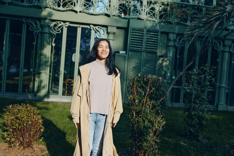Woman outdoor coat casual smile gate garden residence daylight sunshine fashion style vibe urban suburban photo shot candid lifestyle setting fabric denim beige outerwear subject summer greenery