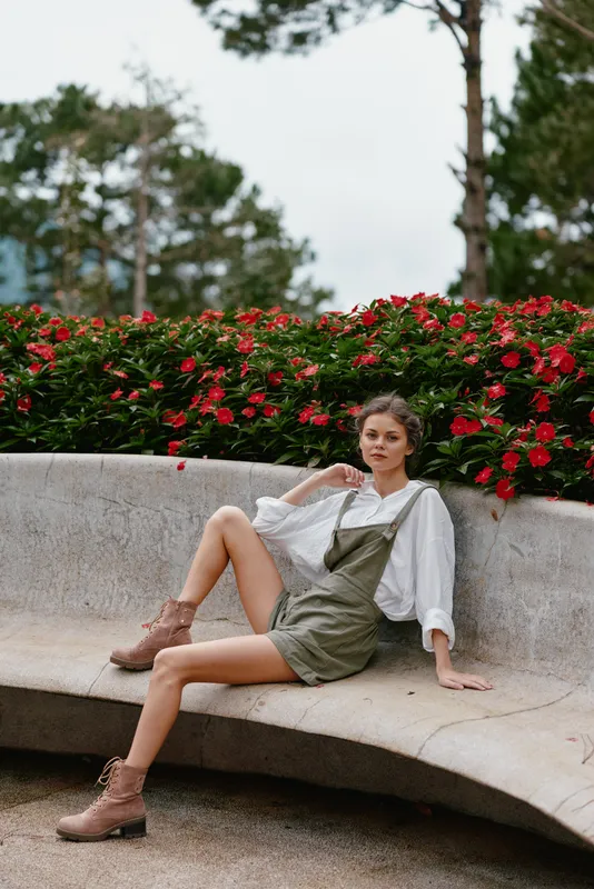 Stylish young woman in casual attire sitting on a stone bench surrounded by vibrant flowers, expressing confidence and relaxation in a natural setting