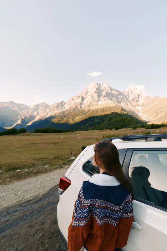 woman and car parked by roadside facing mountains landscape at sunset, travel roadtrip scene with nature meadow and horizon, peaceful outdoor adventure and scenic journey