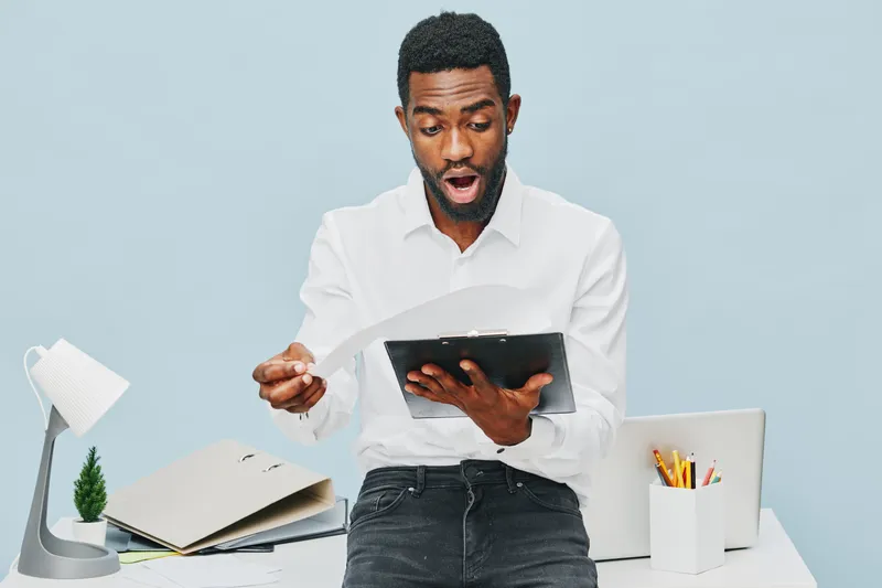 Surprised man with dark skin in white shirt holding tablet device over light blue background, demonstrating shock or amazement, modern workspace with desk, books, and stationery, studio shot. People e