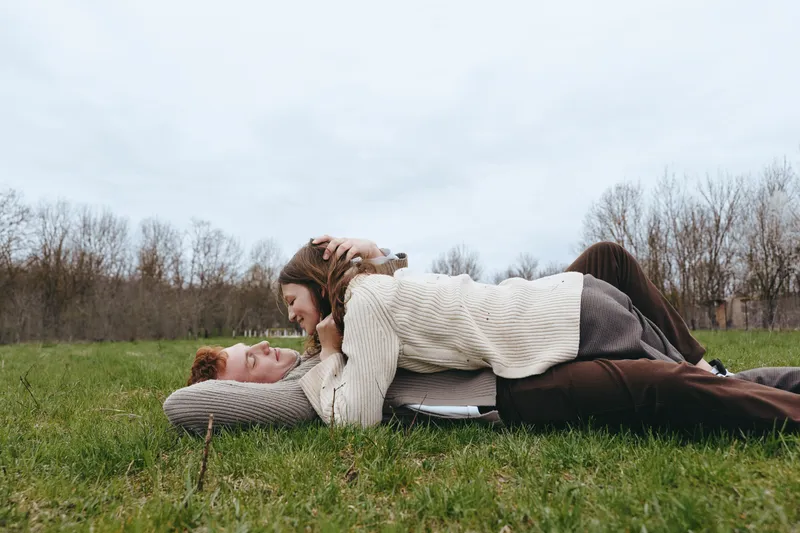 Two people lie on a grassy field sharing a playful cuddle under a cloudy sky, wearing cozy sweaters, embracing in a relaxed pose, enjoying fresh air. This lifestyle moment highlights warmth,