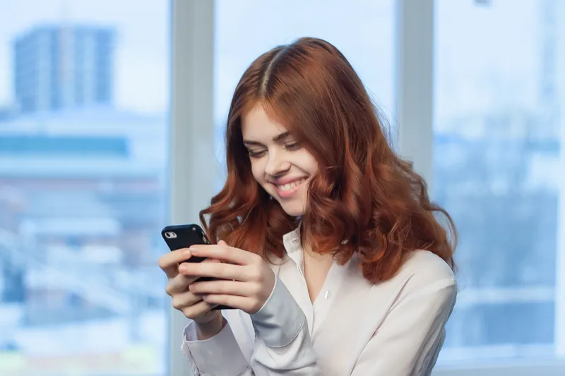 Рыжеволосая девушка улыбается, глядя в смартфон у окна