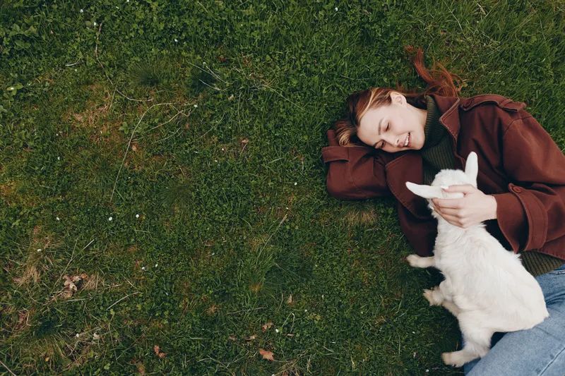 Cheerful outdoor moment of a woman lying on grass, cuddling a white goat kid, warm sunlight, relaxed posture, soft breeze, natural colors, candid lifestyle scene, gentle affection